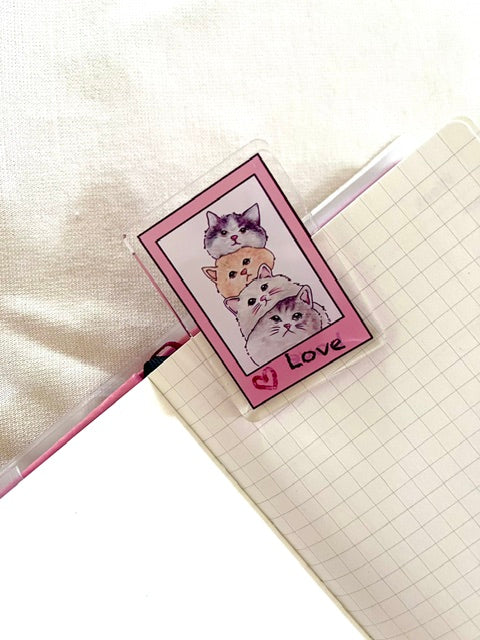 A clear acrylic paper clip featuring a pink border and an illustration of three stacked cats by simple and soft studio
